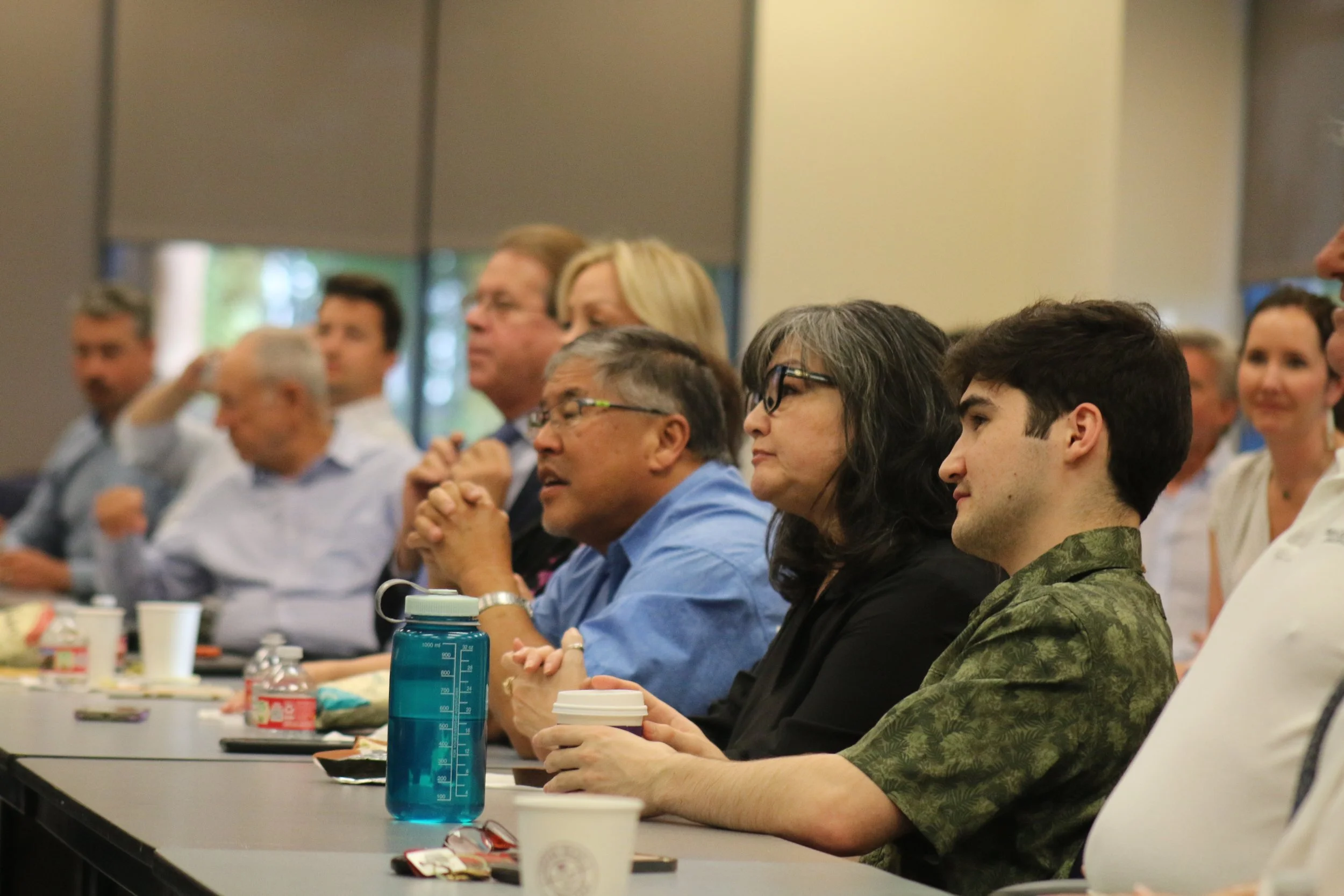 People sitting at a conference table listening attentively, with drinks and snacks in front of them.