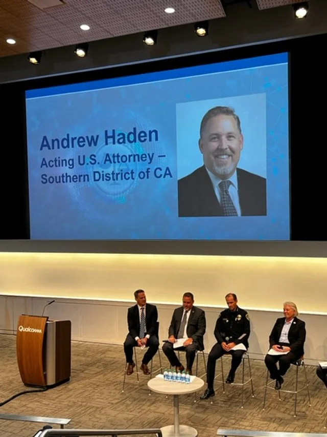 A large auditorium with a screen displaying the name Andrew Haden, Acting U.S. Attorney for the Southern District of California. The screen also shows a photo of Andrew Haden. There are four people sitting on stage and a podium with a laptop in front