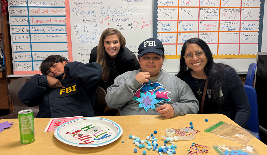 Three women and two children sitting at a table in a classroom, with a whiteboard in the background. The children wear FBI hats, and one child has an FBI jacket. The table has craft supplies, a 'FELV Loved' plate, and decorated paper.