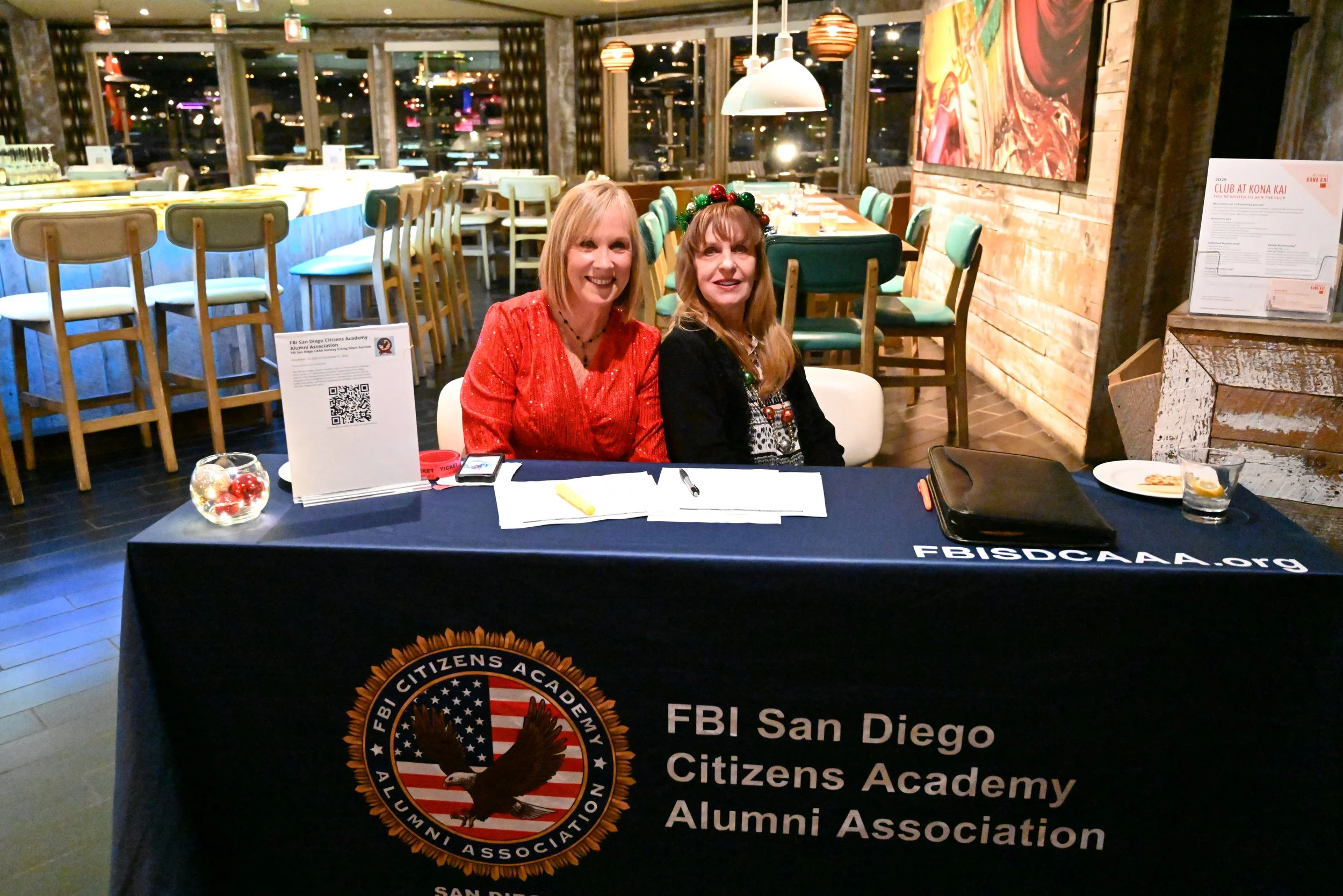 Two women sitting at a table with a black cloth displaying the logo for FBI San Diego Citizens Academy Alumni Association, inside a restaurant or event space with wooden walls, art on the wall, and green chairs in the background.