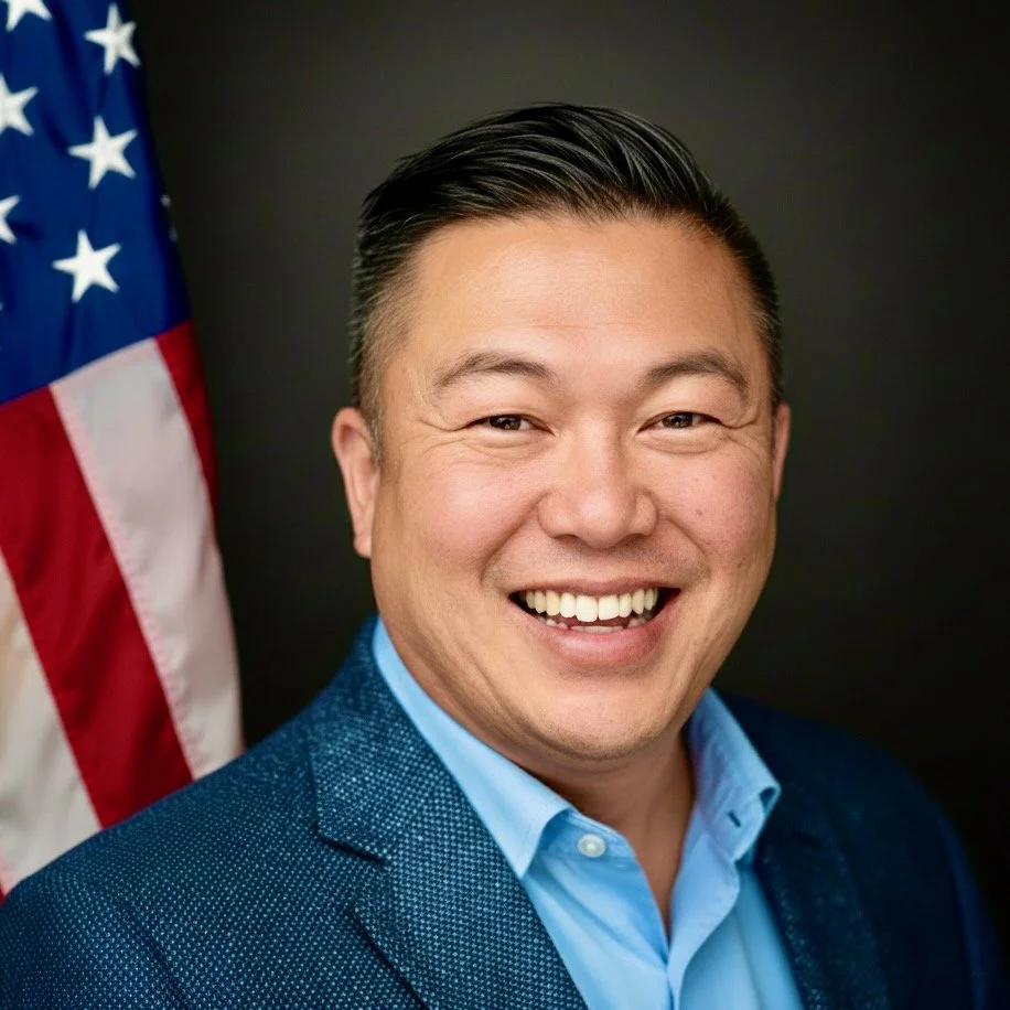 Portrait of a smiling man in a suit and dress shirt, standing in front of an American flag.