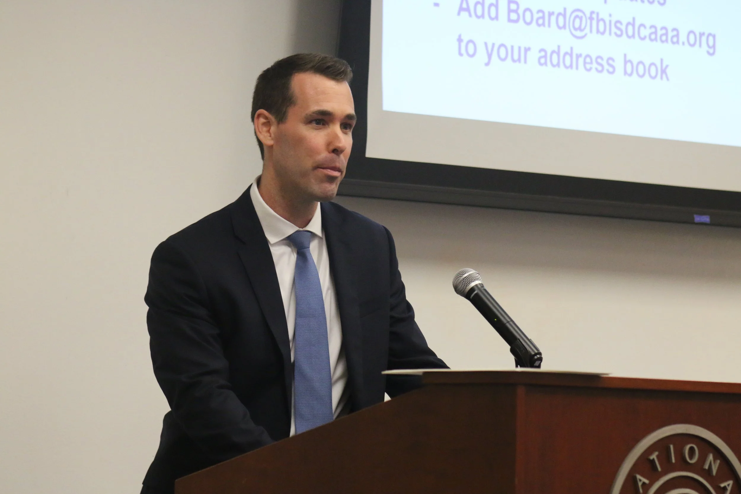 A man in a dark suit and light blue tie stands behind a podium at a conference or meeting, speaking into a microphone. A screen behind him displays instructions related to adding an email address to an address book.