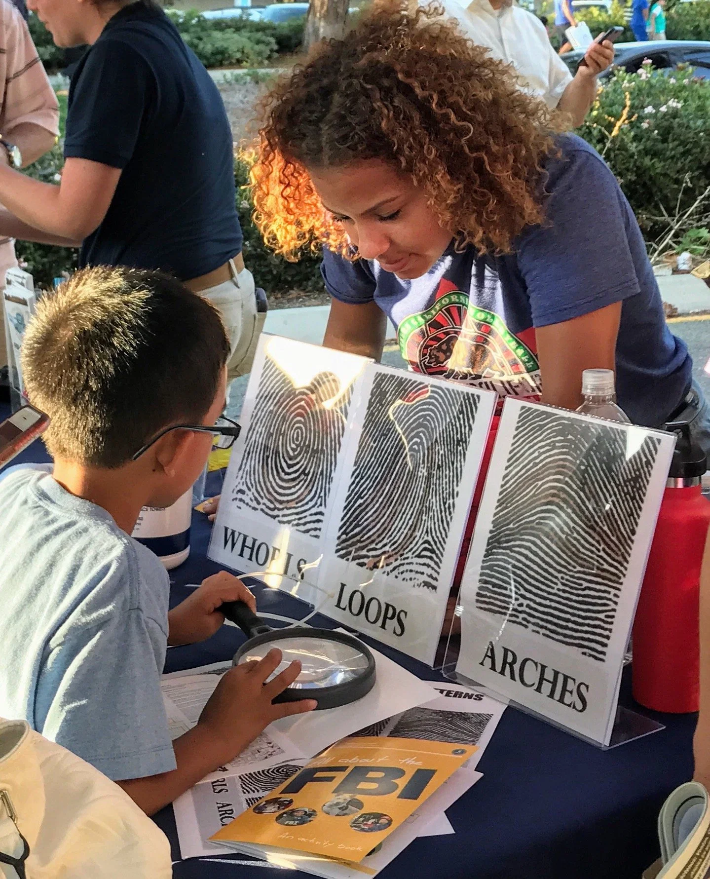 A young boy and a woman at a table examining fingerprint images labeled 'Whorls,' 'Loops,' and 'Arches' during an event about the FBI, with other people in the background.