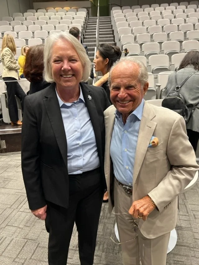 Two elderly individuals, a woman and a man, dressed in business attire, smiling and posing for a photo in a large auditorium or conference hall with empty seats in the background.
