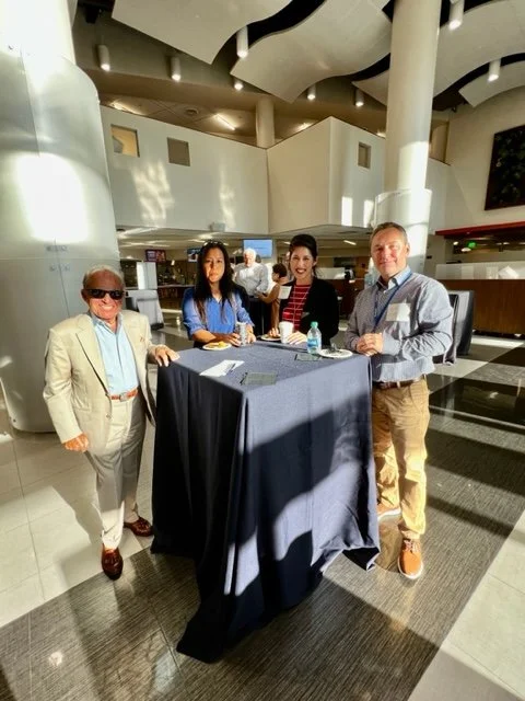 Four people gathered around a high table in a modern indoor space, possibly an airport or conference center, with a bright ceiling and curved architectural elements, engaged in conversation.