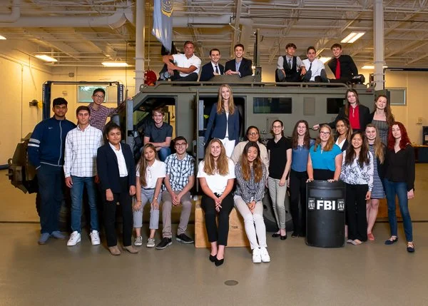 Group of students and FBI agents posing with an armored vehicle inside a warehouse or industrial space.
