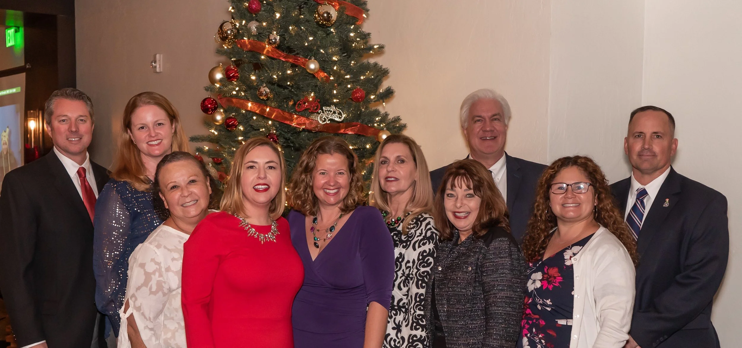 Group of ten people in front of a decorated Christmas tree, smiling at a holiday gathering.