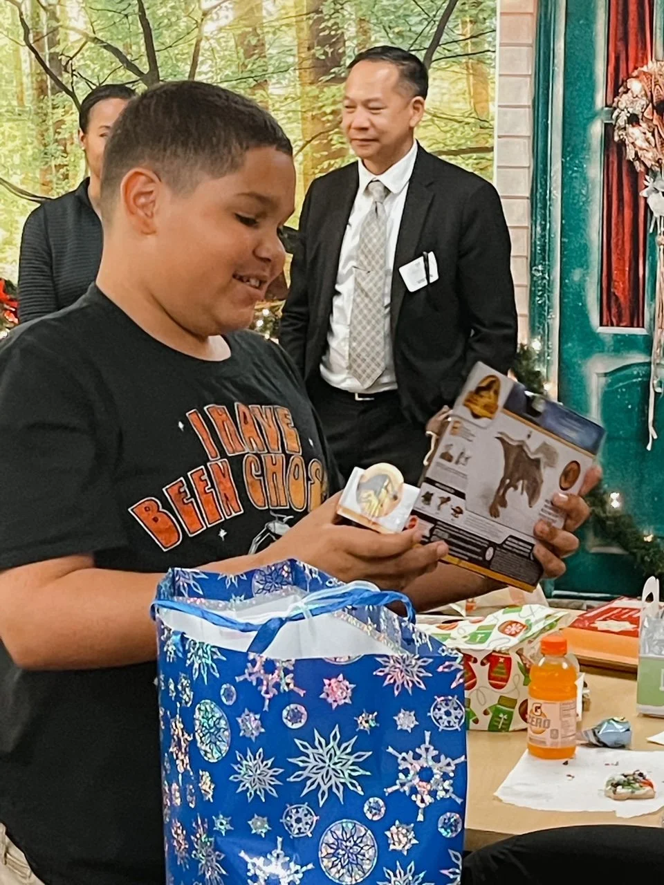 A boy opening a gift at a Christmas party, with a decorated table and adults in the background.