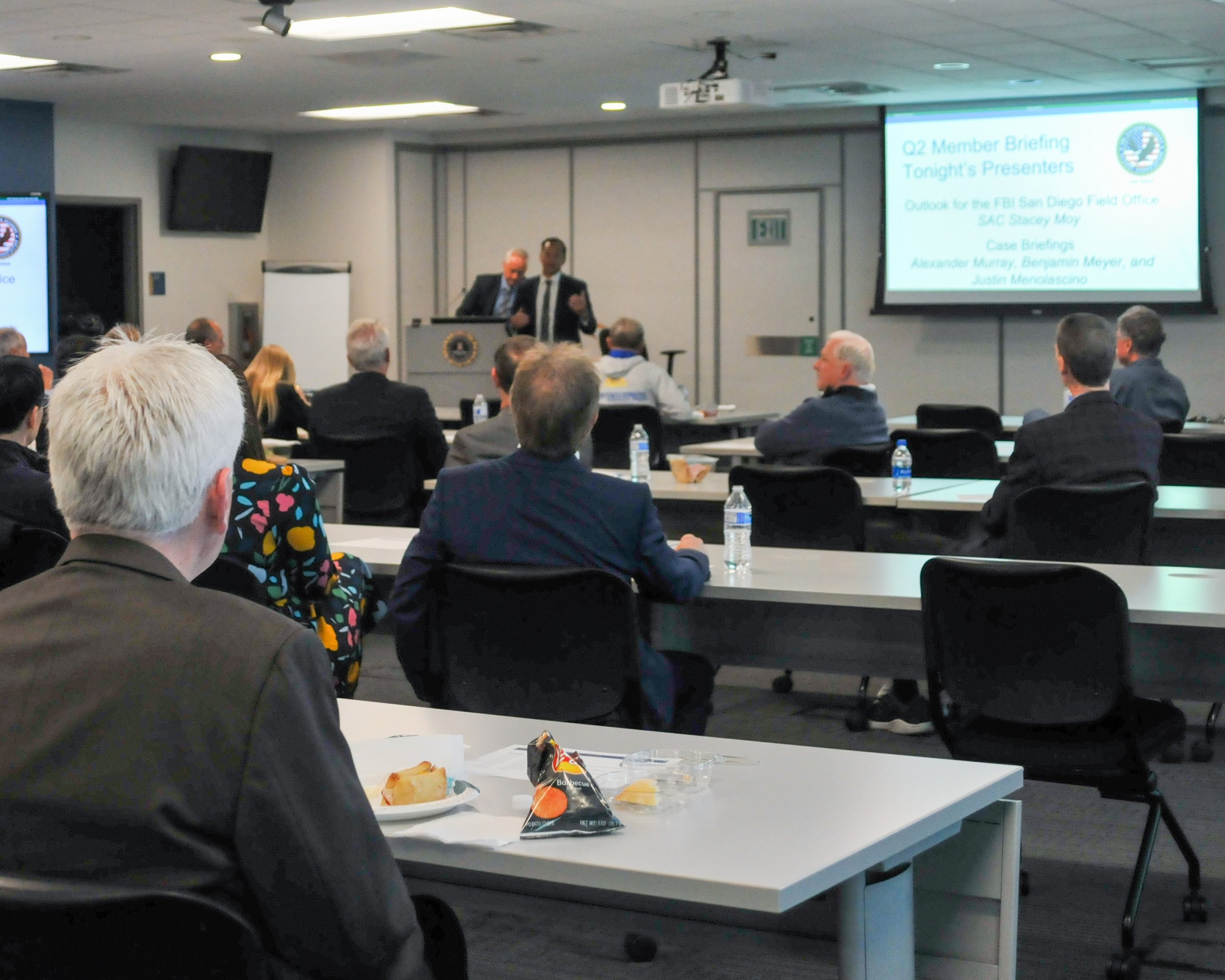 A conference room with attendees seated at tables, listening to a speaker at the front. A presentation slide is visible on the right screen, and water bottles are on the tables.