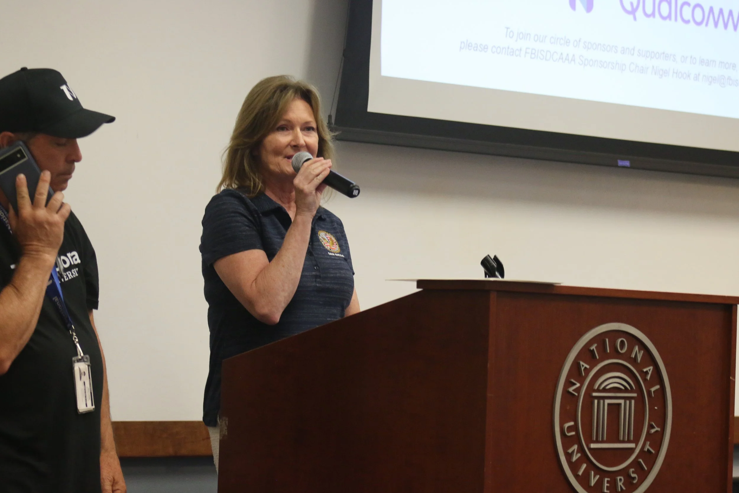 A woman speaking into a microphone at a podium with the logo of an educational institution, with a man beside her talking on a cellphone. A large screen with information about Qualcomm is in the background.