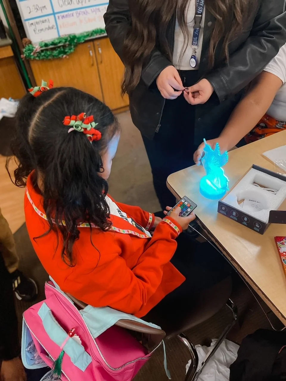 A girl with dark curly hair, wearing orange pajamas and holiday-themed hair accessories, is sitting at a table, using a remote control to operate a small light-up unicorn figurine. Another person, standing, is holding an object near the unicorn. Ther