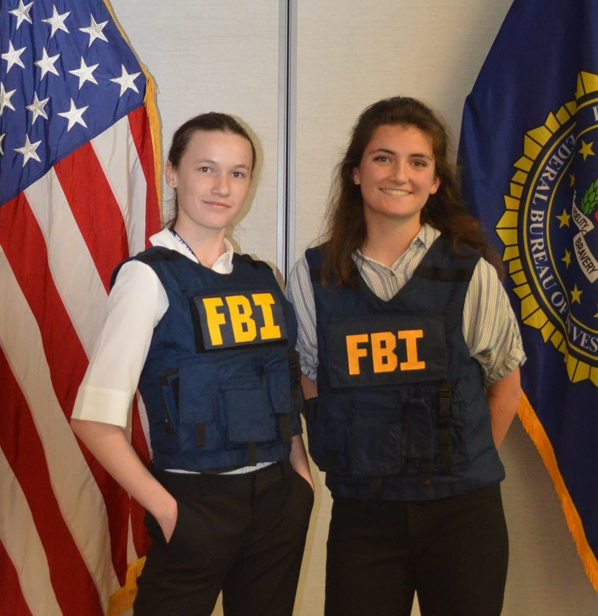 Two women in FBI tactical vests stand in front of American and FBI flags at a formal event.