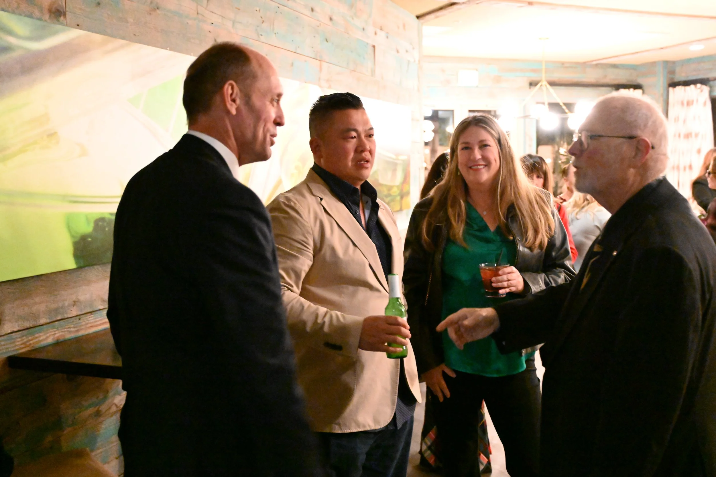 Four adults standing in a social gathering, talking and drinking in a warmly lit room with wood-paneled walls and abstract art.