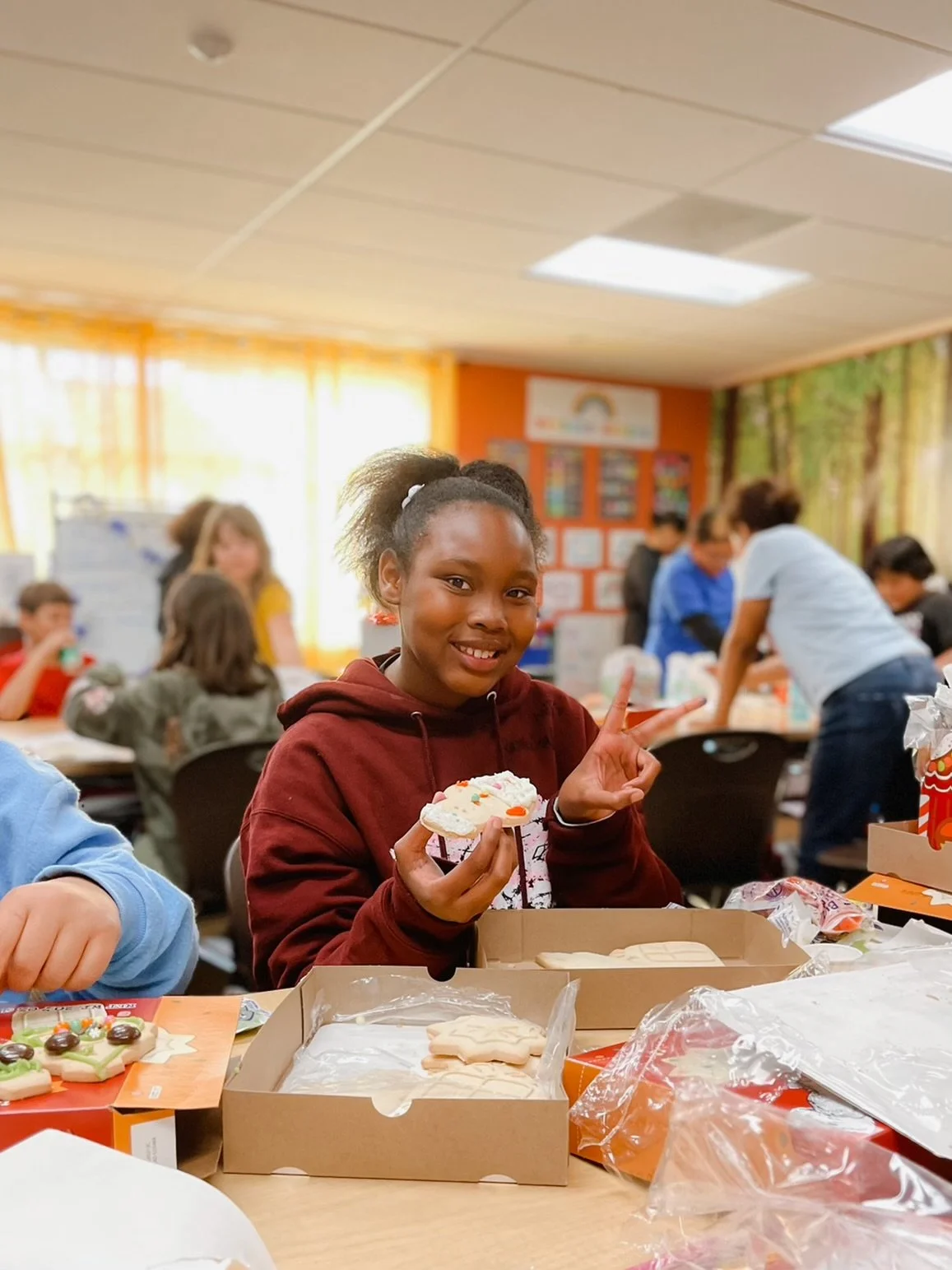 A young girl with a ponytail holding a decorated cookie and making a peace sign, sitting at a table with Christmas cookies and boxes in a festive classroom with other children and adults in the background.