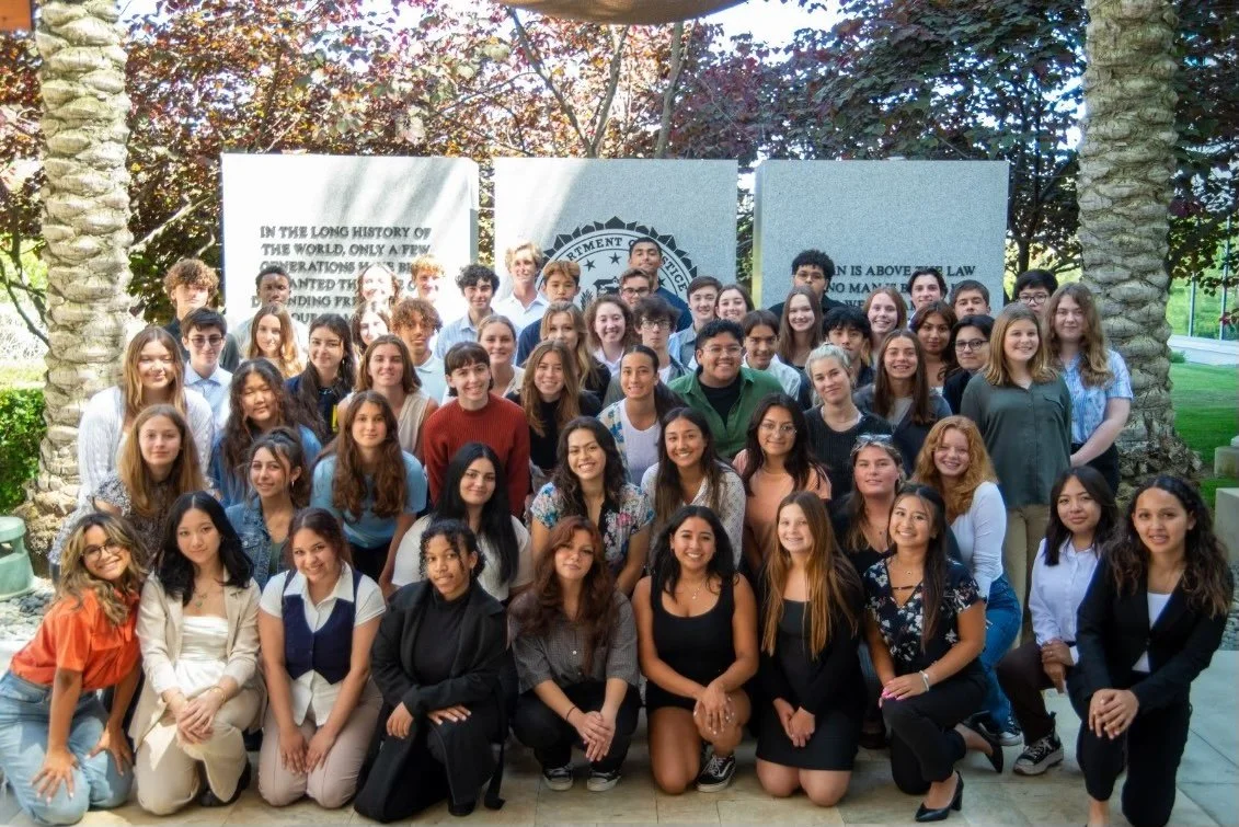 Group of diverse students and teachers gathered outdoors in front of a memorial with flags and trees.