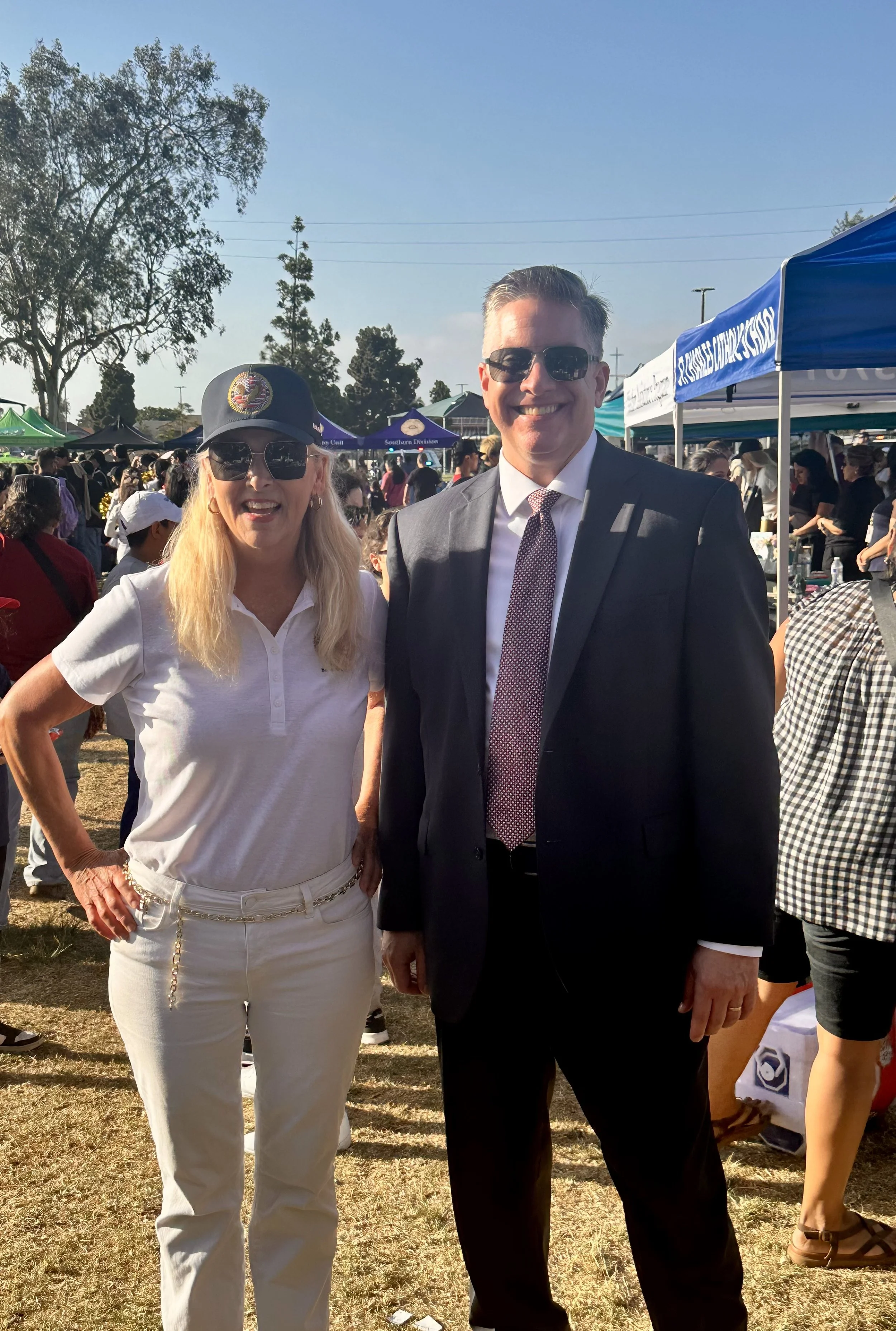 A woman and a man stand together outdoors at a crowded event, both wearing sunglasses. The woman is dressed in a white polo shirt, white pants, and a navy cap, smiling with her hand on her hip. The man is in a dark suit with a white shirt and patterned tie, also smiling.