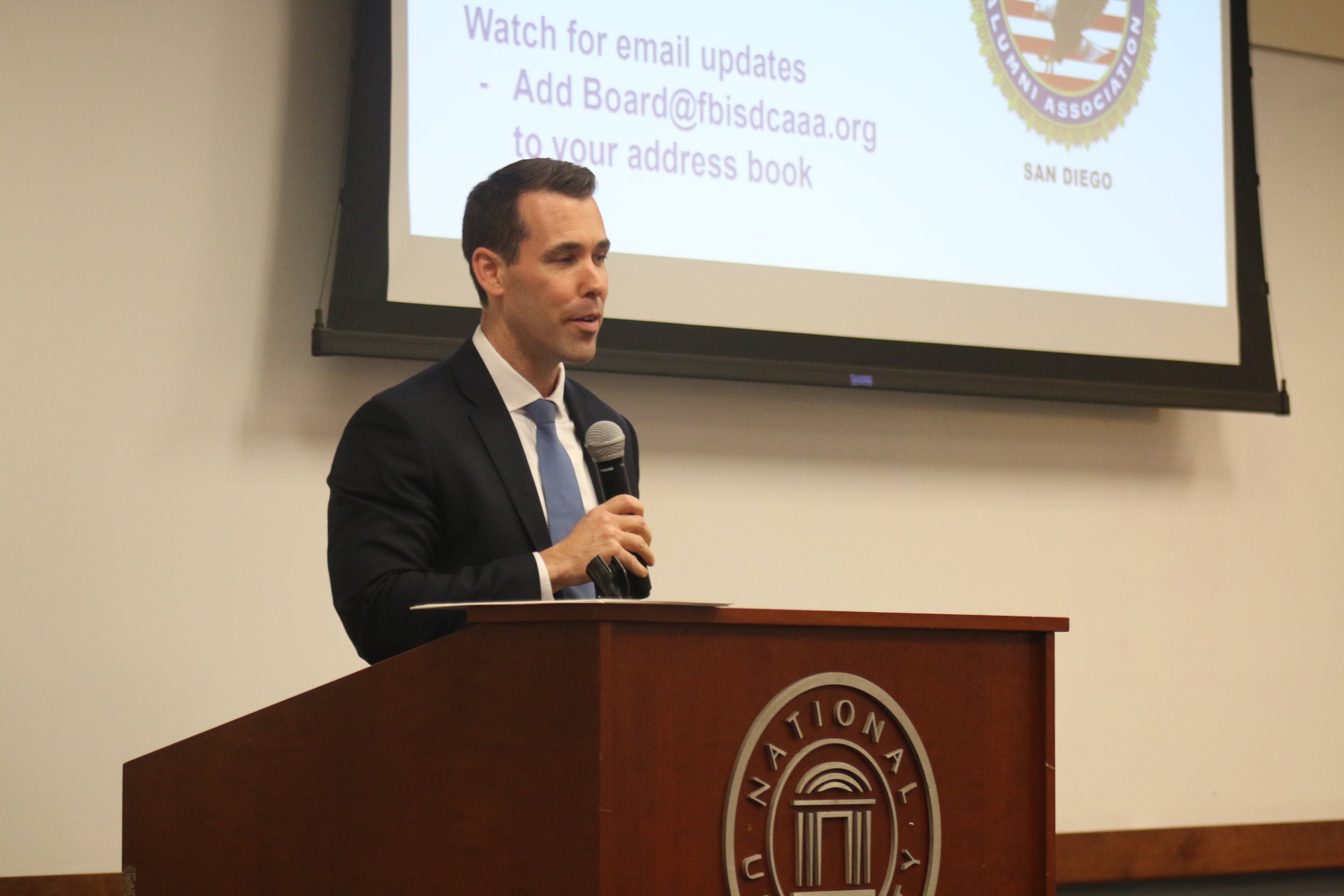 A man in a black suit and light blue tie standing at a podium with the logo of the National University of San Diego, speaking into a microphone. A presentation slide with text and a logo is displayed on a screen behind him.