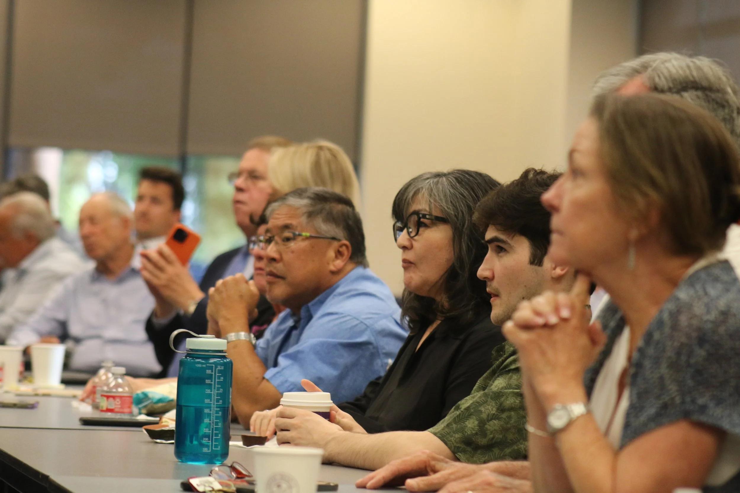 People seated at a conference table listening attentively, with various items like water bottles, cups, and snacks in front of them.