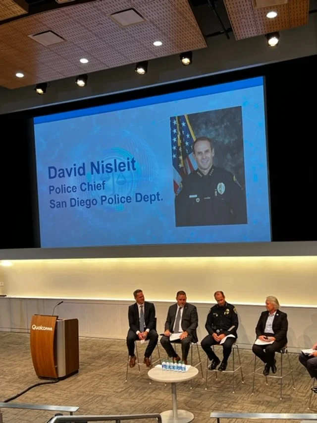 A presentation in a conference room showing a slide with a photo of Police Chief David Niseit of the San Diego Police Department. The slide includes his name and title, with the American flag behind his picture. Four men are seated on stage, and a po