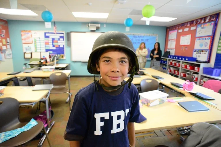 Young boy wearing a firefighter helmet in a classroom with desks, chairs, and school decorations.