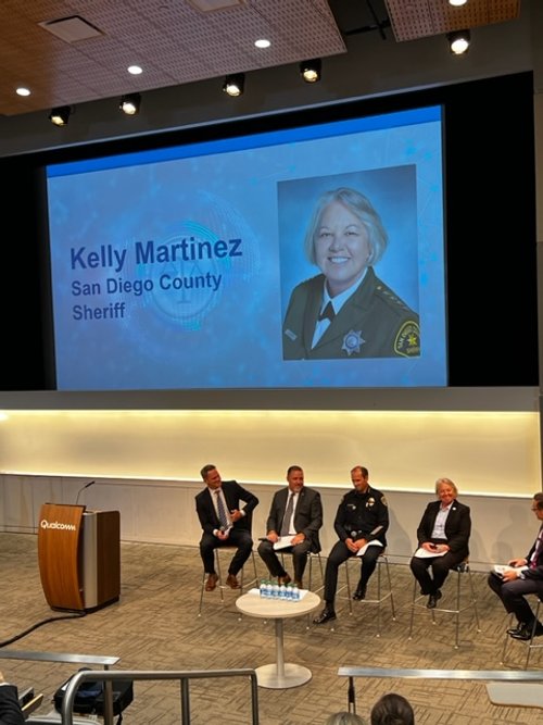 Panel discussion with five men sitting on stage in front of a large screen displaying a photo of Kelly Martinez, a sheriff from San Diego County, in a dark uniform with a badge. One man is standing at a podium labeled Qualcomm.