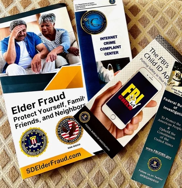 A collection of printed materials and a smartphone on a table. The materials include a brochure about elder fraud, a card from the Internet Crime Complaint Center, a pamphlet about the FBI's Child ID App, and other federal law enforcement information