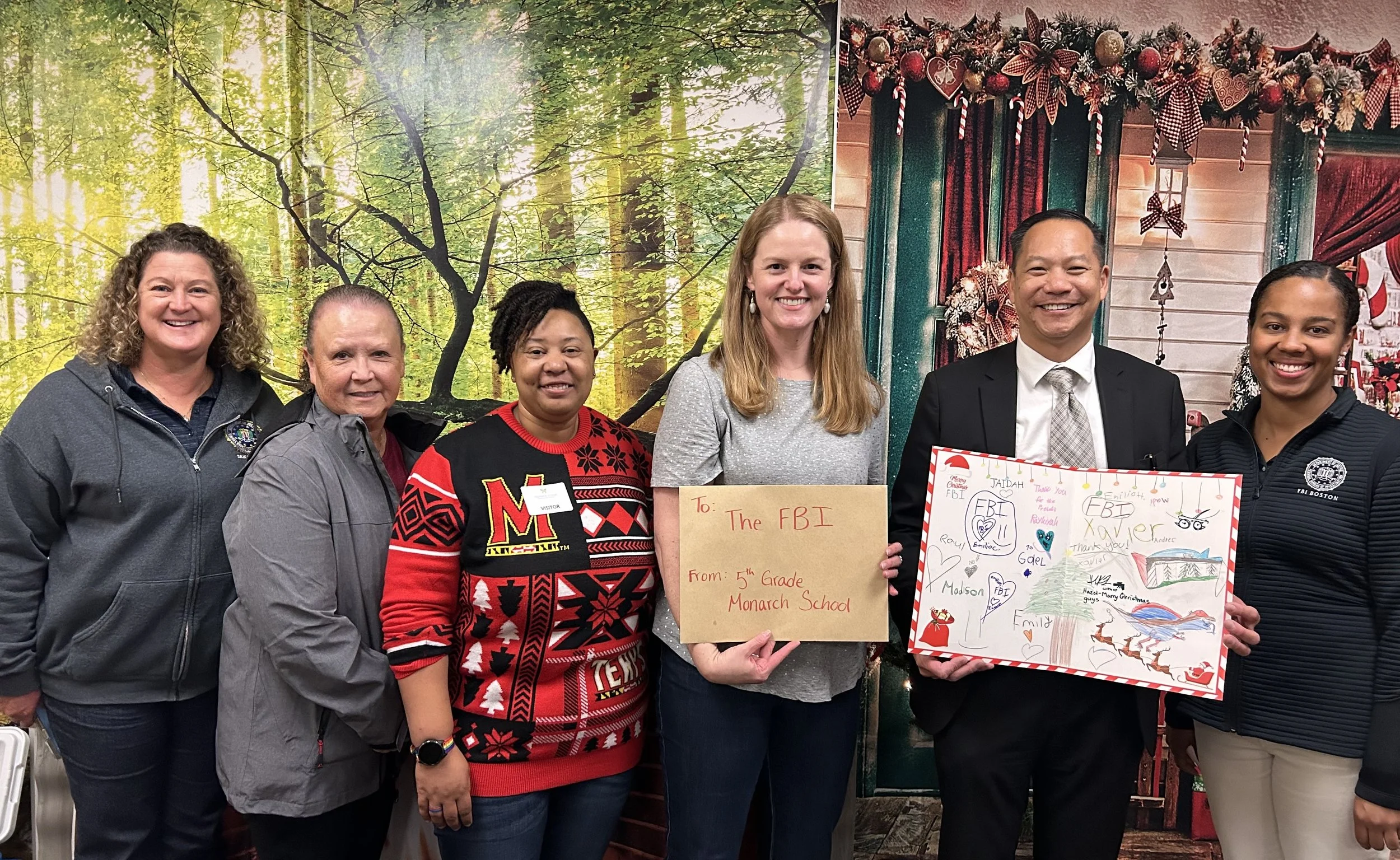 Six people stand together smiling in front of a festive background with trees and Christmas decorations. Two individuals in the center are holding signs with messages to the FBI, from a 5th grade class, during a holiday event.