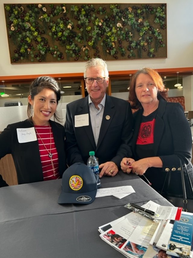 Three people, two women and one man, posing together at a table in a restaurant or conference setting. The woman on the left has long dark hair and is wearing a black blazer and red striped shirt. The man in the middle has gray hair, glasses, and is 