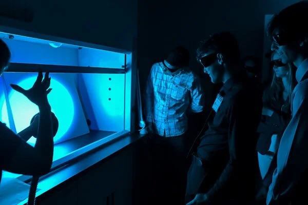People observing a large, illuminated, blue, circular device or exhibit in a dark room.
