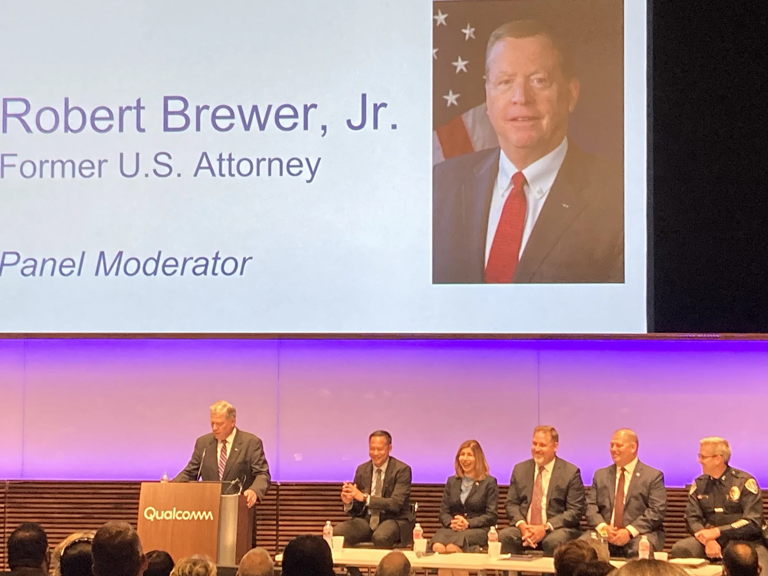 Panel with six people, one man at a podium labeled Qualcomm, and a large screen displaying a photo and information about Robert Brewer Jr., a former U.S. Attorney, as the moderator of the panel.