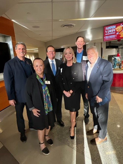 Six people standing together in an indoor setting, smiling for a group photo. There are three men and three women, dressed in business and casual attire. A red and pink sign in the background reads 'Pumpkin & Spice' and 'Treat Yourself'.