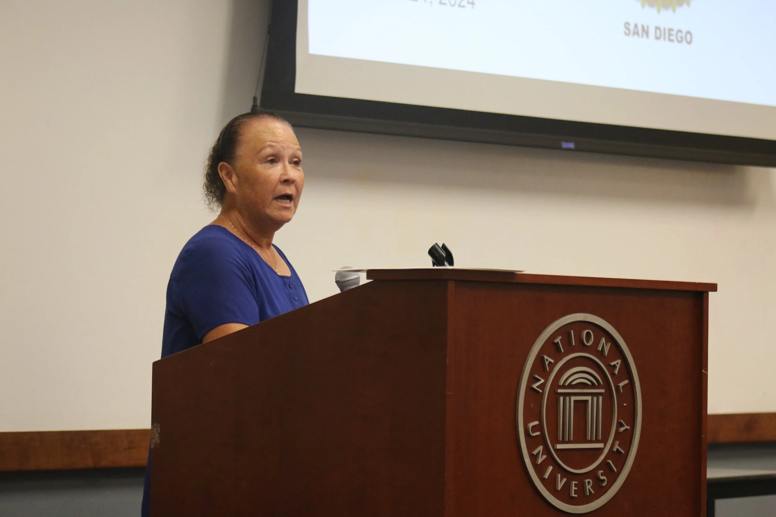 A middle-aged woman speaking at a podium with a university seal, wearing a blue top, in a classroom or lecture hall at National University.