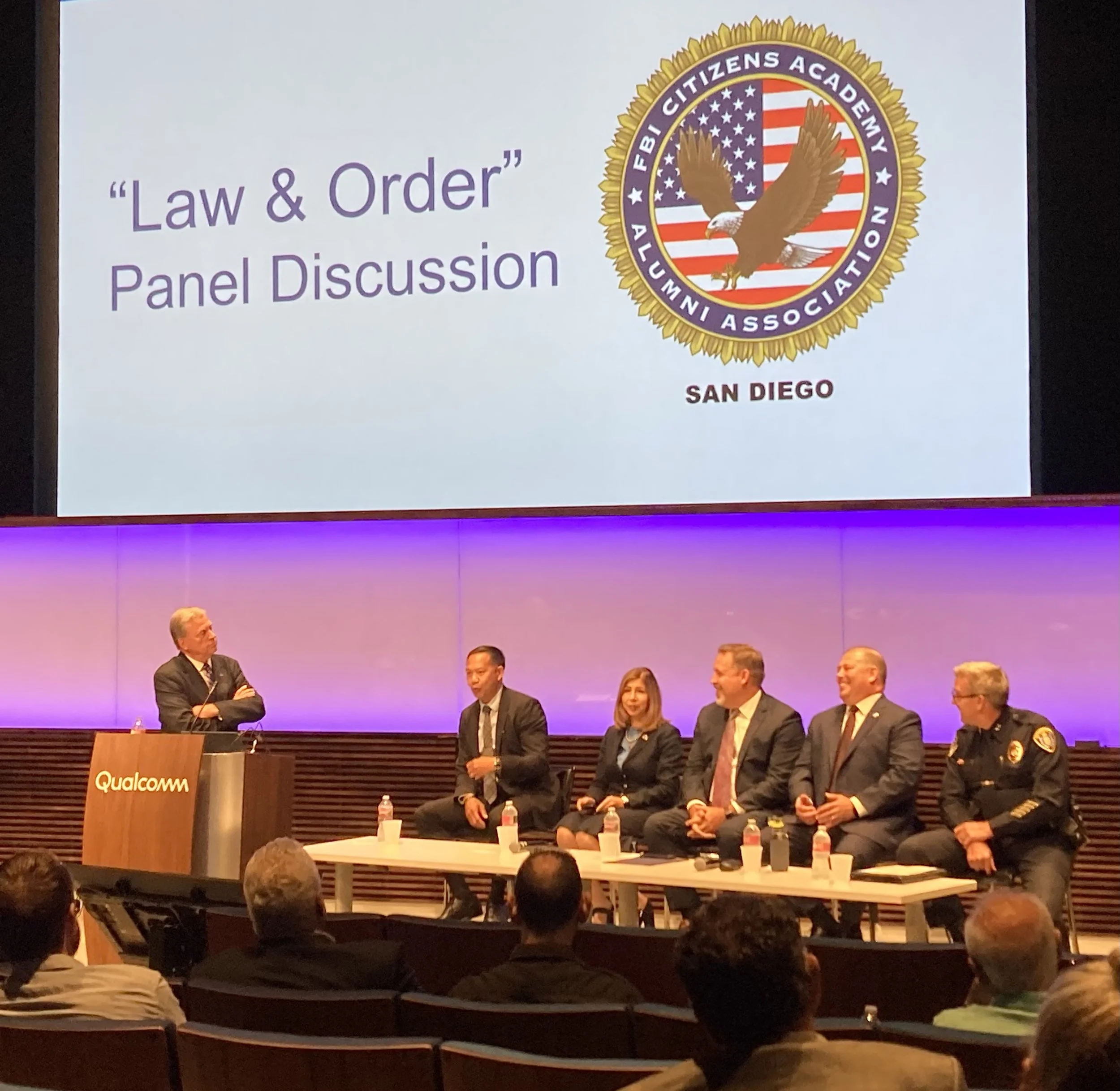 Panel discussion on 'Law & Order' at Qualcomm auditorium, with five speakers seated at a table on stage, and a large screen displaying the event title, logo, and location, San Diego.