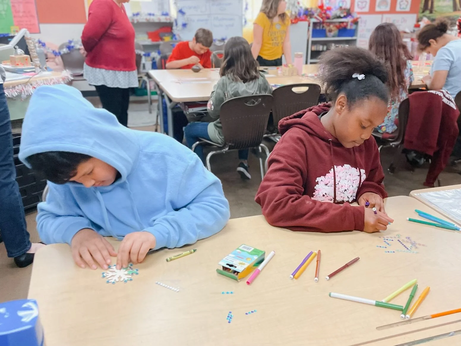 Children working on holiday craft projects at a classroom table, decorating snowflakes with colorful stickers and markers.