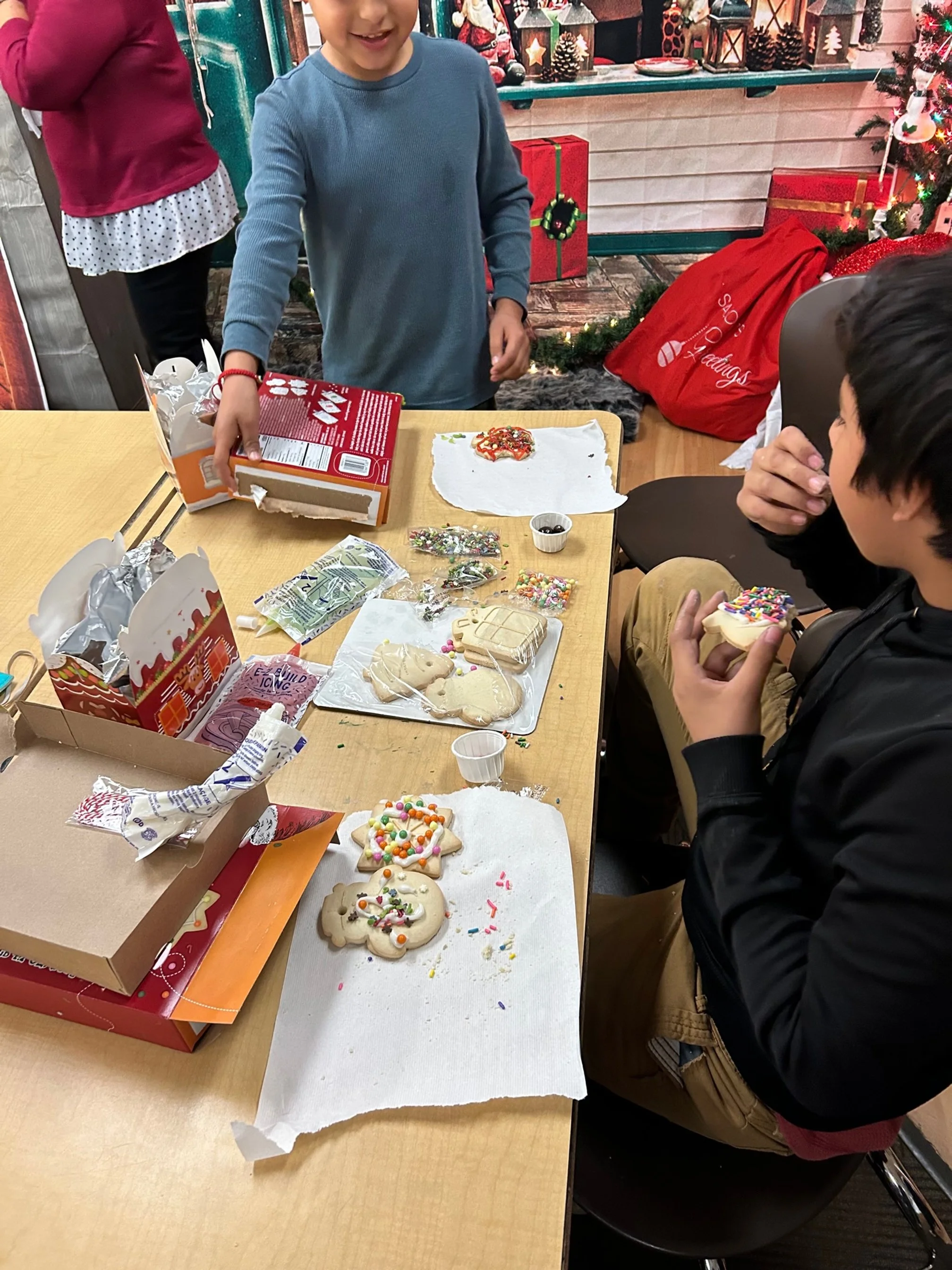 Children decorating Christmas cookies at a table with holiday-themed cookies, sprinkles, and icing in a festively decorated room.