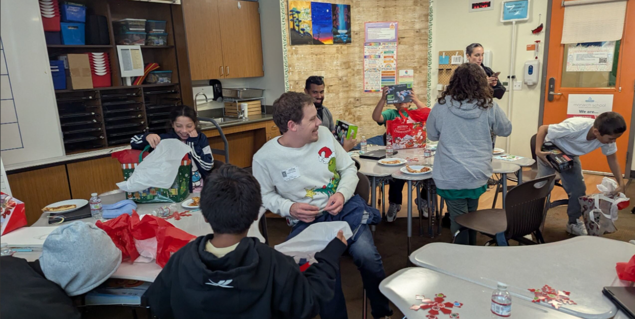 Group of children and adults gathered in a classroom for a holiday celebration, opening gifts and wearing festive attire.
