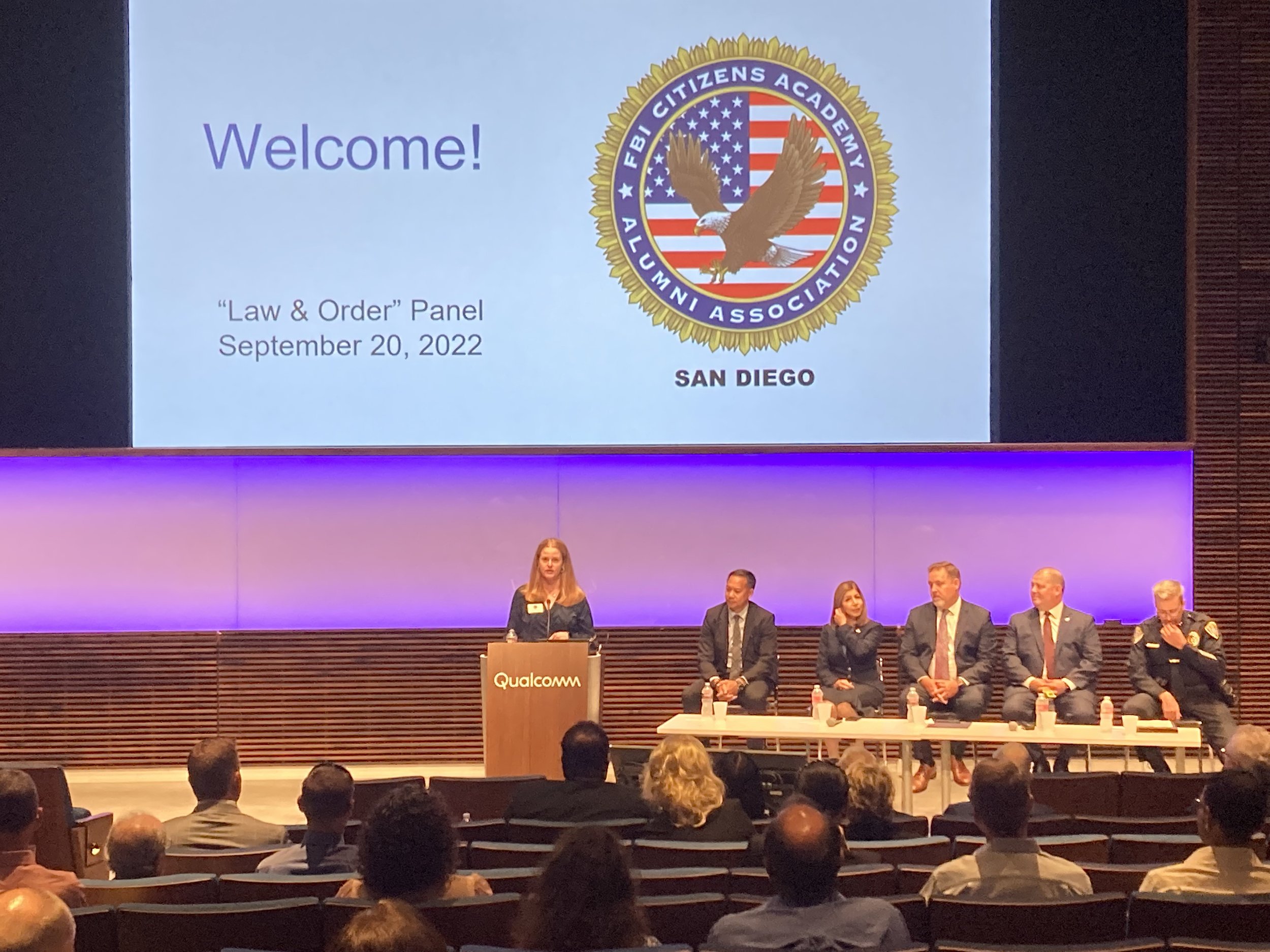 A panel discussion at a conference with a large screen displaying a welcome message for the 'Law & Order' panel on September 20, 2022, organized by the FBI Citizens Academy Alumni Association in San Diego. Five people sit at a table on stage, and one woman is speaking at a podium labeled Qualcomm.