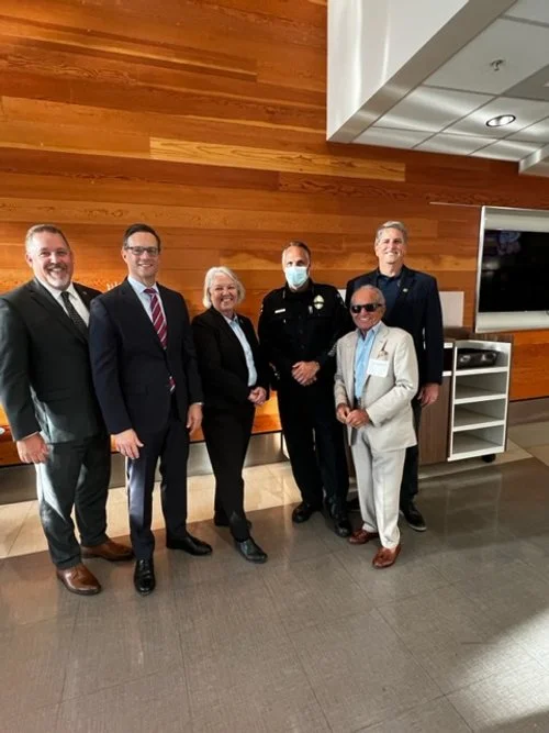Six people standing together indoors, five men and one woman, with a wooden wall and a TV in the background. One man is in a police uniform, and another is wearing sunglasses and a light-colored suit.