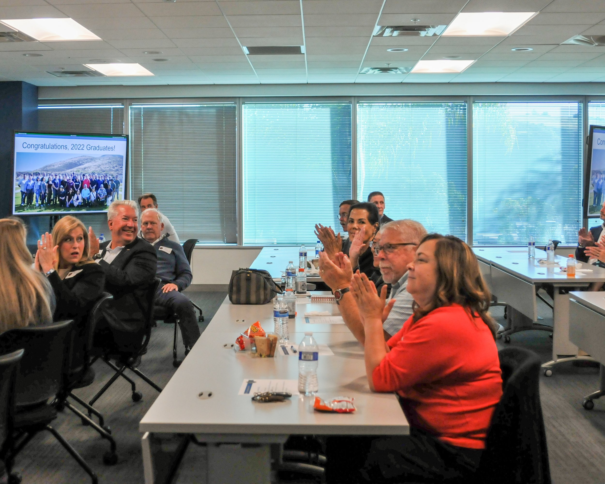 People seated at tables in a conference room, attending a graduation celebration, with a screen displaying "Congratulations, 2022 Graduates!" showing a group photo outside.