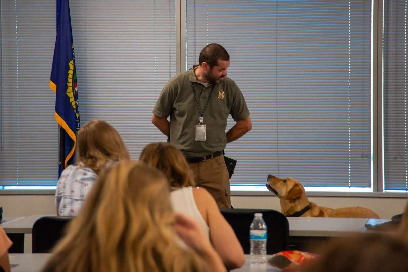 A man in a gray shirt with a badge and lanyard stands in a classroom or seminar room, looking at a dog lying on a table. Several women are seated at tables with water bottles, listening. The room has large windows with closed blinds and a flag in the