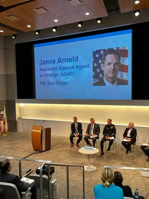 Panel discussion at a conference with four men seated on stage, a large screen behind them displaying a presentation slide about Jamie Arnold, an Assistant Special Agent in Charge at FBI San Diego. The slide includes a photo of Jamie Arnold with an A