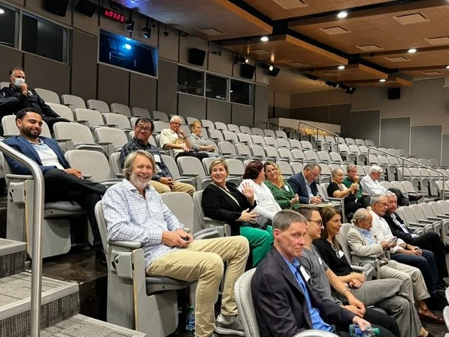 A group of people sitting in a movie theater or auditorium, spaced out with some wearing masks, some smiling or waving, with empty seats around them.