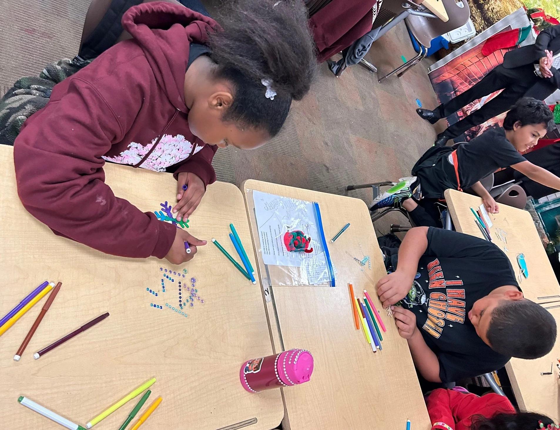 Child creating a craft project with colorful beads on a wooden table in a classroom decorated for Christmas.