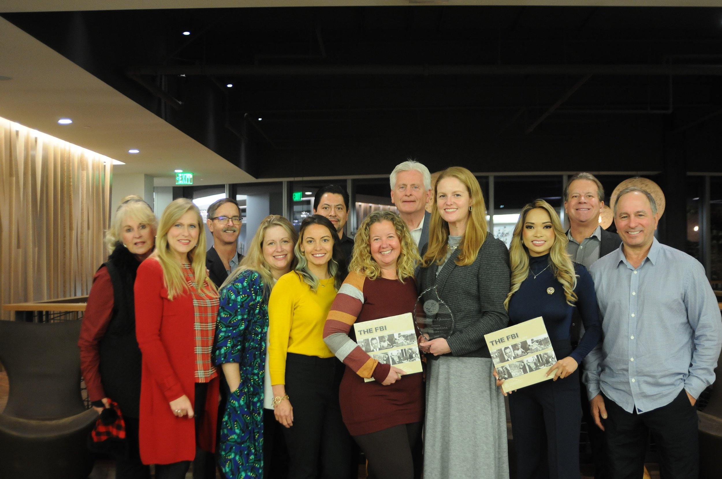 A group of diverse adults smiling and posing for a photo at an indoor event, with two women holding awards and certificates labeled 'The FBI'.