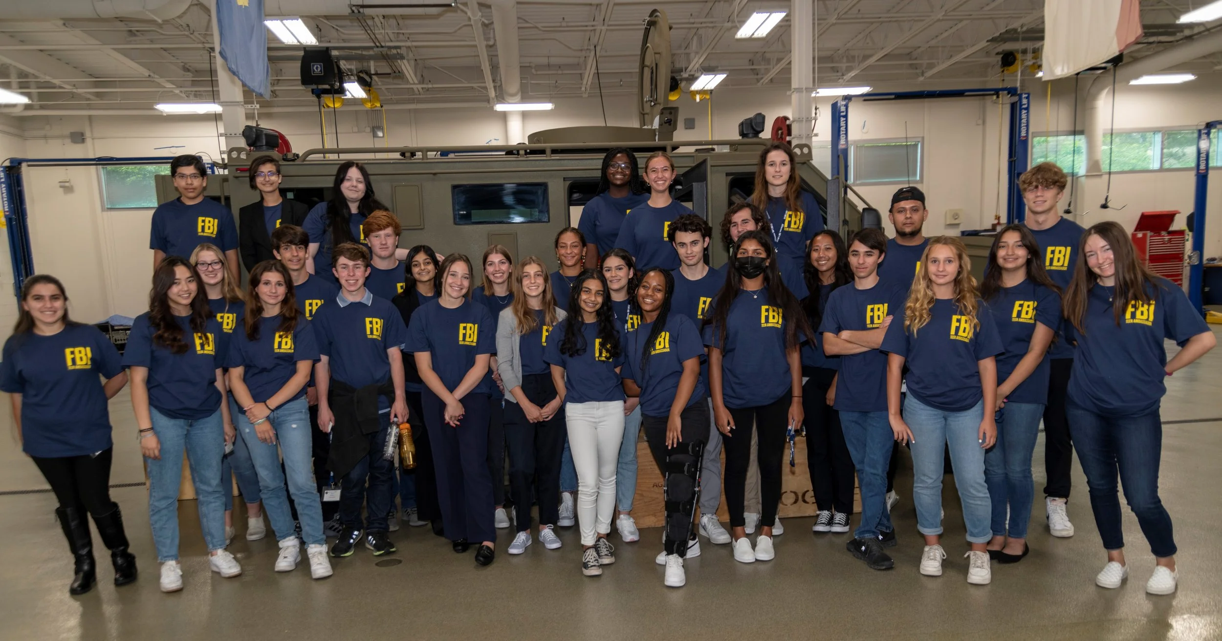 Group of young people wearing FBI T-shirts standing inside a workshop with machinery and an armored vehicle in the background.