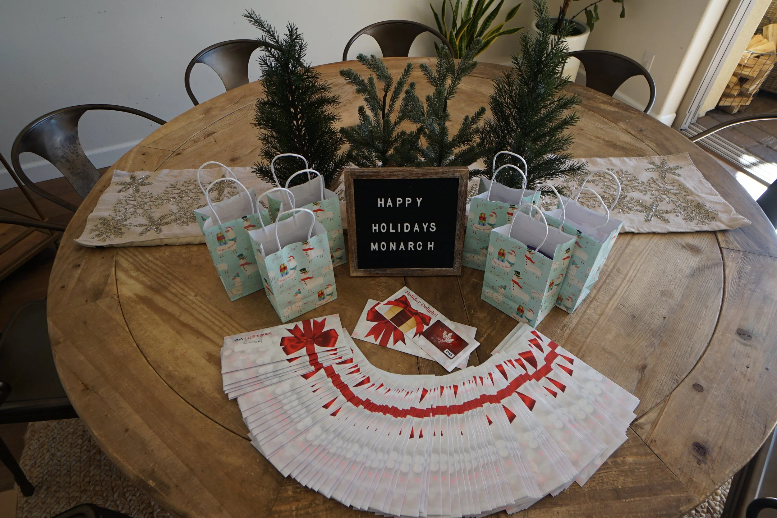 A wooden dining table decorated with holiday items, including a small pine tree centerpiece, a black framed sign that reads 'HAPPY HOLIDAYS MONARCH', several gift bags with holiday designs, a fan of red and white holiday cards, and some wrapped gift 
