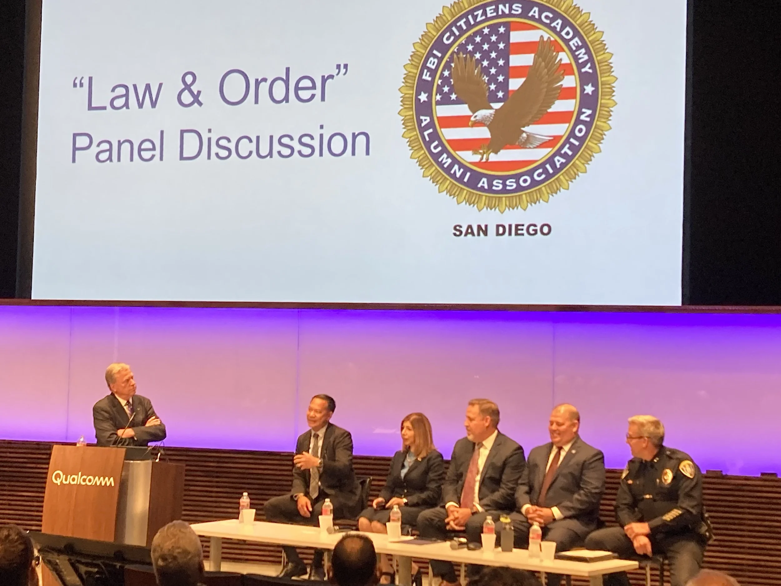 A panel discussion titled 'Law & Order' at Qualcomm, with six people sitting on stage and one person standing at a podium, in front of a large screen displaying the FBI Citizens Academy Alumni Association logo and the words 'San Diego.'