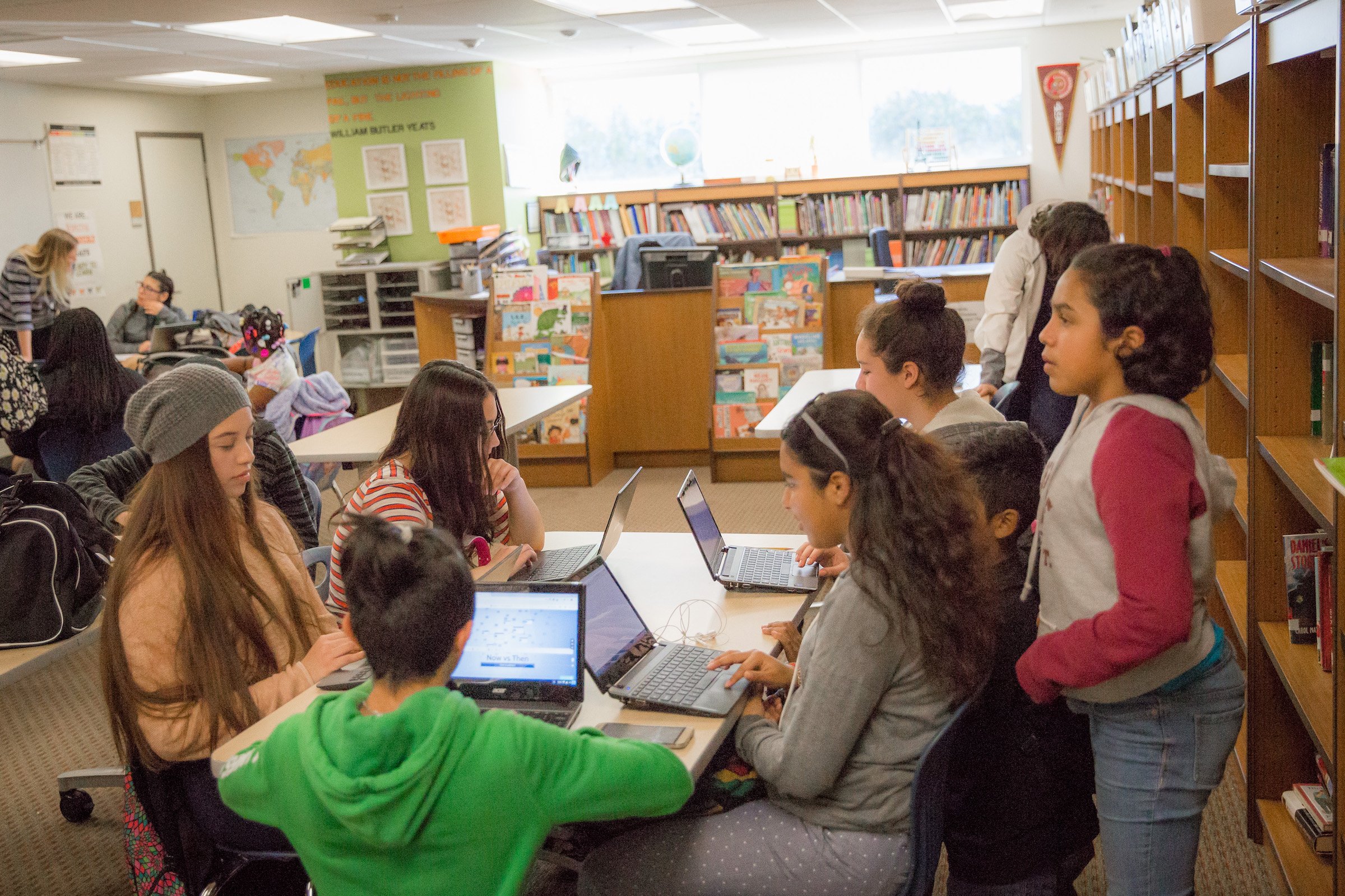 Children working on laptops in a classroom filled with bookshelves.