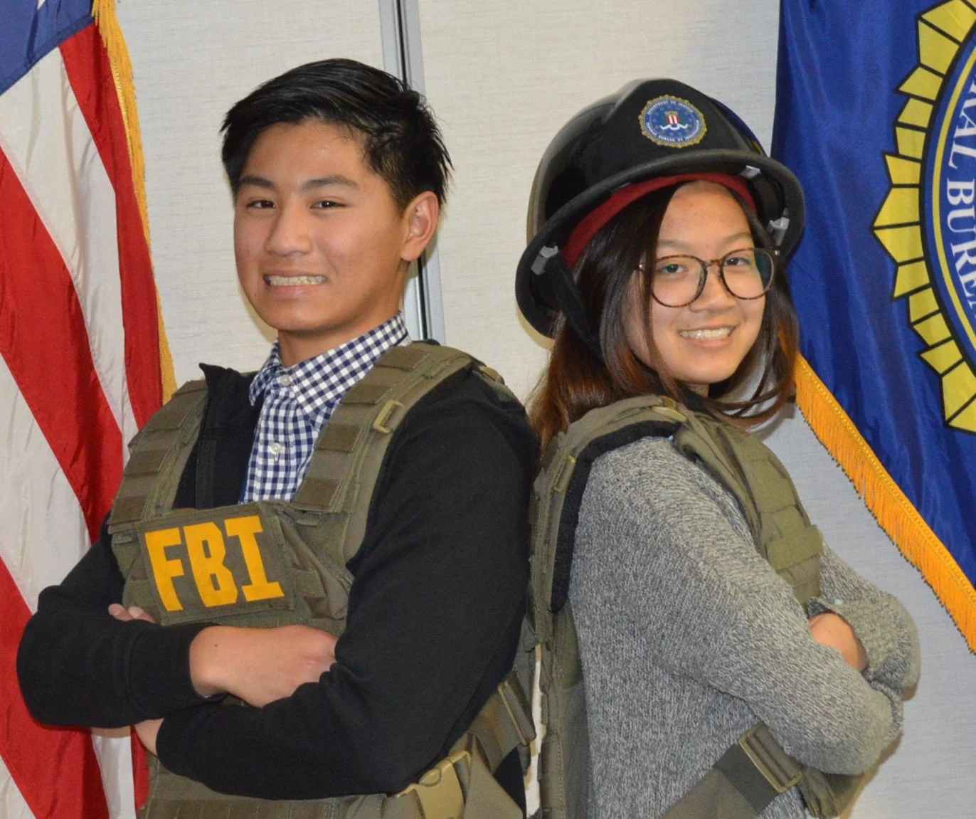 Two young adults, one male and one female, standing back to back with arms crossed, wearing tactical vests and gear, with flags in the background.