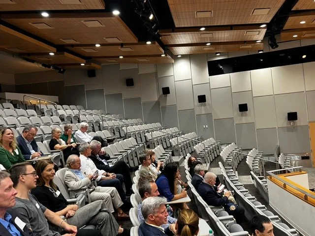 Audience seated in a conference or lecture hall with tiered seating and wood-paneled ceiling